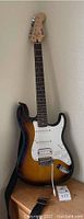 Full view of Fender Squier Bullet Strat guitar standing, showing sunburst body with white pickguard and attached shoulder strap.