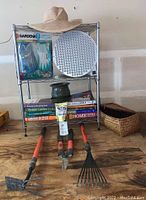 Shelf with gardening gloves in original package, straw hat on top, green ceramic pot on middle shelf, several gardening books stacked, and gardening hand tools in front: rake, cultivator, pruning shears.