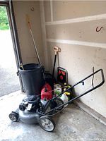 Lawnmower with garbage can, garden rakes, gas can, plastic containers and garden hand tools positioned in a garage corner.