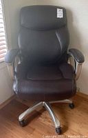 Front view of TrueWellness dark brown leather-like office chair showing high backrest, padded armrests, and metal base with casters