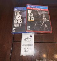 Photo of two sealed PS4 game cases: The Last of Us Part II in blue case and The Last of Us Remastered in red PlayStation Hits case, placed on a wooden surface with auction label 051.