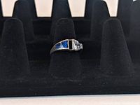 Silver ring with black opal inlay stones arranged in a row next to a clear pear-shaped stone, displayed on a black ring holder.