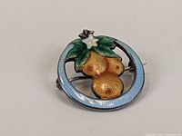 Front angle showing the brooch's guilloche enamel circular frame and detailed orange cluster with green leaves and white flower.