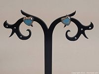 Front view of the pair of silver heart-shaped stud earrings with green stones mounted on decorative black display stand.