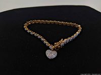 Bracelet fully visible showing alternating gold plated and silver links with clear stones, heart charm attached, laying on black background.