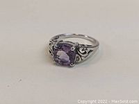 Front angled view of sterling silver ring with faceted amethyst-colored stone and decorative scrollwork band.