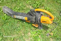 Side view of the WORX electric blower mulcher vacuum on grass showing orange-black body and WORX branding on nozzle.