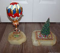 Two Ron Lee porcelain figurines on a floor: a hot air balloon and a Christmas tree, showing size and relative scale