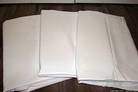 Three folded white polyester tablecloths laid side by side on a wooden floor, showing their size and texture.