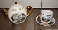 Photo showing the Thomas Kinkade ceramic teapot and matching cup and saucer with painted cottage scenes and gold accents.