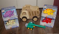 Six toddler push toys grouped together: 4 cartoon triceratops dinosaur toys in boxes and loose, a large unfinished wooden dump truck, and a green wooden crane truck on wooden floor.