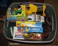 Overhead photo of a tote filled with various children's toys including a boxed Junior Racer Tractor, Marvel Rescue Jet playset, and toy trucks.