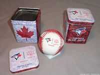 Photo showing two square tins and one baseball out of the tin with lid removed to reveal foam padding inside.
