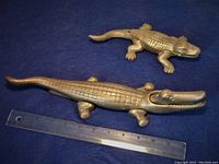 Two solid brass alligator or crocodile figures placed next to a ruler for size reference on a blue background.