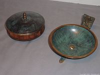 Two metal decorative bowls on a grey surface, one covered with wooden sides and metal lid, the other an open bowl with green patina interior and ornate feet