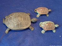 Three turtle figurines placed on a blue cloth background showing the large brass turtle alongside two smaller copper-tone turtles.