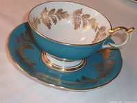 Blue porcelain cup with gold leaf design inside and outside, curved gold-accented handle, paired with matching saucer decorated similarly in blue with gold leaf and trim.