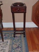 Full front view showing the wooden smoker's stand with single drawer and circular ashtray on top, positioned on a rug beside furniture.