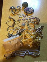 Photo showing assorted jewelry pieces including bracelets, necklaces, rings, a compact mirror, and a heart-shaped ring box on a yellow surface.
