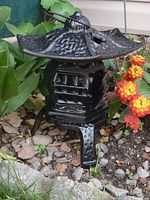 Black cast iron pagoda lantern outdoors among plants, showing the overall form and surroundings.