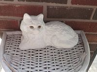 White concrete cat sculpture with painted facial features, displayed on a wicker surface against brick background.
