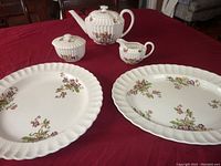 Full set of serving pieces on red tablecloth showing teapot, sugar bowl, creamer, and two platters with floral patterns and basketweave rims.
