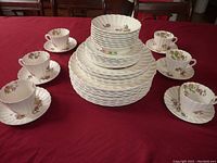 Photo showing six teacups each paired with a saucer arranged around stacks of plates and bowls featuring the Wicker Lane pattern.