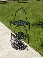 Full view of the black metal folding corner shelf standing outdoors on pavement with grass background.