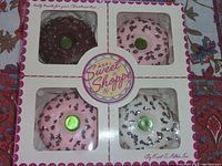 Boxed set of four Kurt S. Adler glass Christmas ornaments shaped like sprinkle donuts, showing front of original packaging with visible branding and donut shapes through box window.