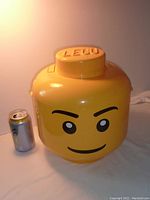 Front view of the large yellow LEGO head container showing the face and LEGO logo stud lid, includes a soda can for size comparison