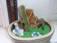 Miniature garden planted in oval glazed ceramic planter with live cactus, miniature pottery chairs, ceramic figurine, decorative turtle, rocks, and artificial grass.