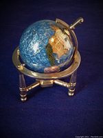 Small size decorative globe with blue enamel surface, gold coordinate lines and multi-colored landmasses, displayed on a polished brass stand with three legs and a circular frame holding the globe.