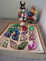 Snowman figurine in background with box of assorted glass ornaments in foreground