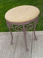 Photo showing full solid metal vanity stool with yellow floral patterned cushioned seat, painted metal base with scrollwork detail, standing outdoors on sidewalk and lawn