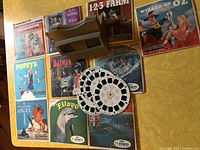Photo shows multiple View-Master reel sets arranged with titles visible including Goldilocks, Wizard of Oz, 1-2-3 Farm, Batman, Popeye, Flipper, and others. Several loose reels are positioned in the center.
