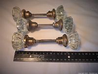 Three pairs of vintage glass door knobs laid flat showing clear faceted glass knobs and brass metal fittings with attached threaded spindles lined up with a ruler for scale.