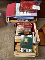 Two cardboard boxes filled with assorted hardcover and paperback books in various conditions, viewed from above with some titles visible.