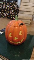 Ceramic pumpkin jar with Jack-o'-lantern face and black spider decoration on top, shown on a dark surface