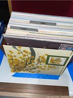 Image showing a box with neatly organized first day issue postcards with tab dividers labeled 1990 and 1991, one postcard with a floral design and commemorative stamp visible.