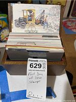 Photo showing a wooden box filled with postcards, a visible card with a vintage truck illustration postmarked 1982, and a tag stating 'First Day Issue Stamps'