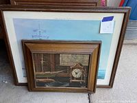 Two framed prints stacked against a wall, larger seascape print behind smaller print of vintage clock and ship model.