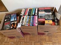 Three open cardboard boxes filled with VHS tapes showing assorted titles