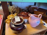 Overview of four vintage ceramic items on wooden shelf: mustard yellow teapot with butterfly designs, maroon covered pot, pink pitcher marked $10, and brown/cream covered pot.