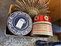 Vintage Syroco jeweled wall clock with a sunburst design partially visible behind assorted vintage cookie tins in a box, showing blue, white/red, and red-lidded tins.
