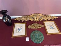 Wide view of lot showing heavy glass vase, large and smaller carved wood gilt door pediments, two framed rose prints, and a vintage green Aztec resin wall hanging arranged against a wall.