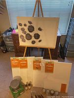 Wooden easel with canvas showing paint spots and four new canvases in packaging, plus some paint jars and mediums on floor
