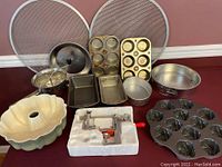 Lot view showing kitchen bakeware and gadgets including muffin pans, bundt pan, loaf pans, pie pans, pizza grates, tomato crusher, and Pampered Chef apple peeler in packaging