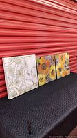 Three floral canvases displayed against a red corrugated background, showing size and colors: white and green canvas with two smaller gray/orange/blue canvases.
