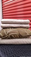 Stacked bedding pieces showing two beige coverlets with scalloped and embroidered designs, one brown and beige botanical toss cushion, all folded.
