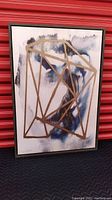 Framed modern abstract art featuring geometric bronze lines over blue and white canvas.